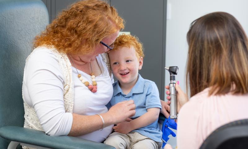 Mother holding her smiling child on her lap as a doctor checks the child’s ear temperature