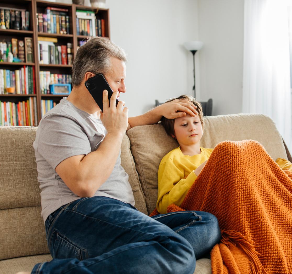Father on a couch talking on the phone while checking his child’s forehead